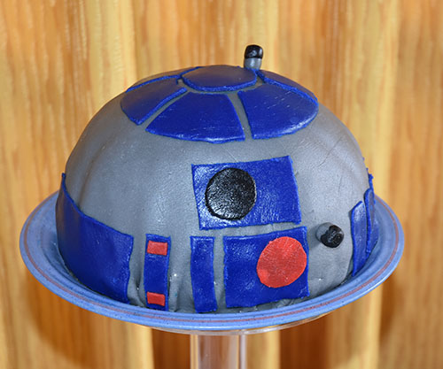 Den tredje kaka var en kopi av R2-D2, og vakte skikkelig jubel fra konfirmanten Jeg laget røre til bløtkakebunn La bakepapir i langpanna, og helt oppi røra, og stekte den i ca 10 minutter, til den var gjennomstekt, og avkjølte den Så tok jeg en bolle og la plast rundt kantene Jeg skar store trekanter av kakebunnen som jeg la langs sidene og bunnen av bollen. Avsluttet med mindre biter som jeg plasserte der det var glipper, og skar vekk eventuelle overlapp. Kantene og bunnen var helt dekt! Så laget jeg jordbærmousse, som jeg skulle ha til fyll i kaka, og Delte jordbær i småbiter, som jeg blandet i moussen. Deretter helte jeg moussen med jordbærbitene oppi kaka, og Lot den stå i kjøleskapet natta over. Morgen etter tok jeg ut kaka, hvelvet den, tok vekk plasten, og Pisket krem, som jeg dekte hele kaka med. Så farget jeg marsipan, og pyntet R2-D2.